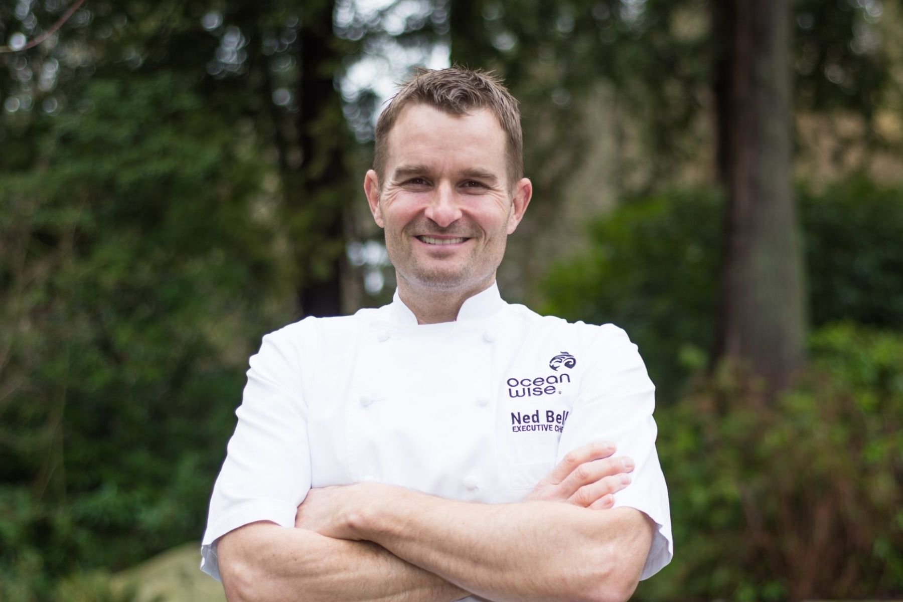 Chef Ned Bell in a white chef jacket, smilling with crossed arms in front of a green forest.