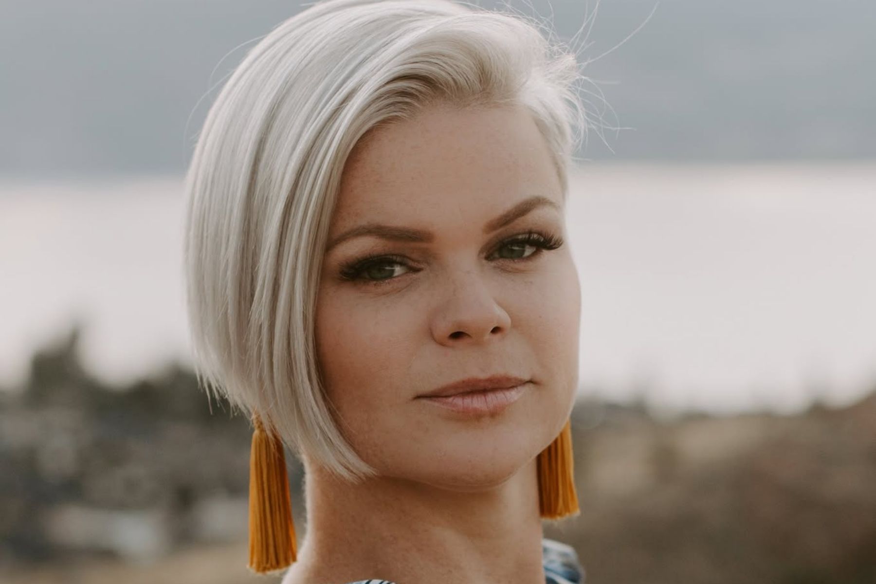 Sara Campbell, with a short platinum blond haircut and dangling orange earrings in front of a lake background.