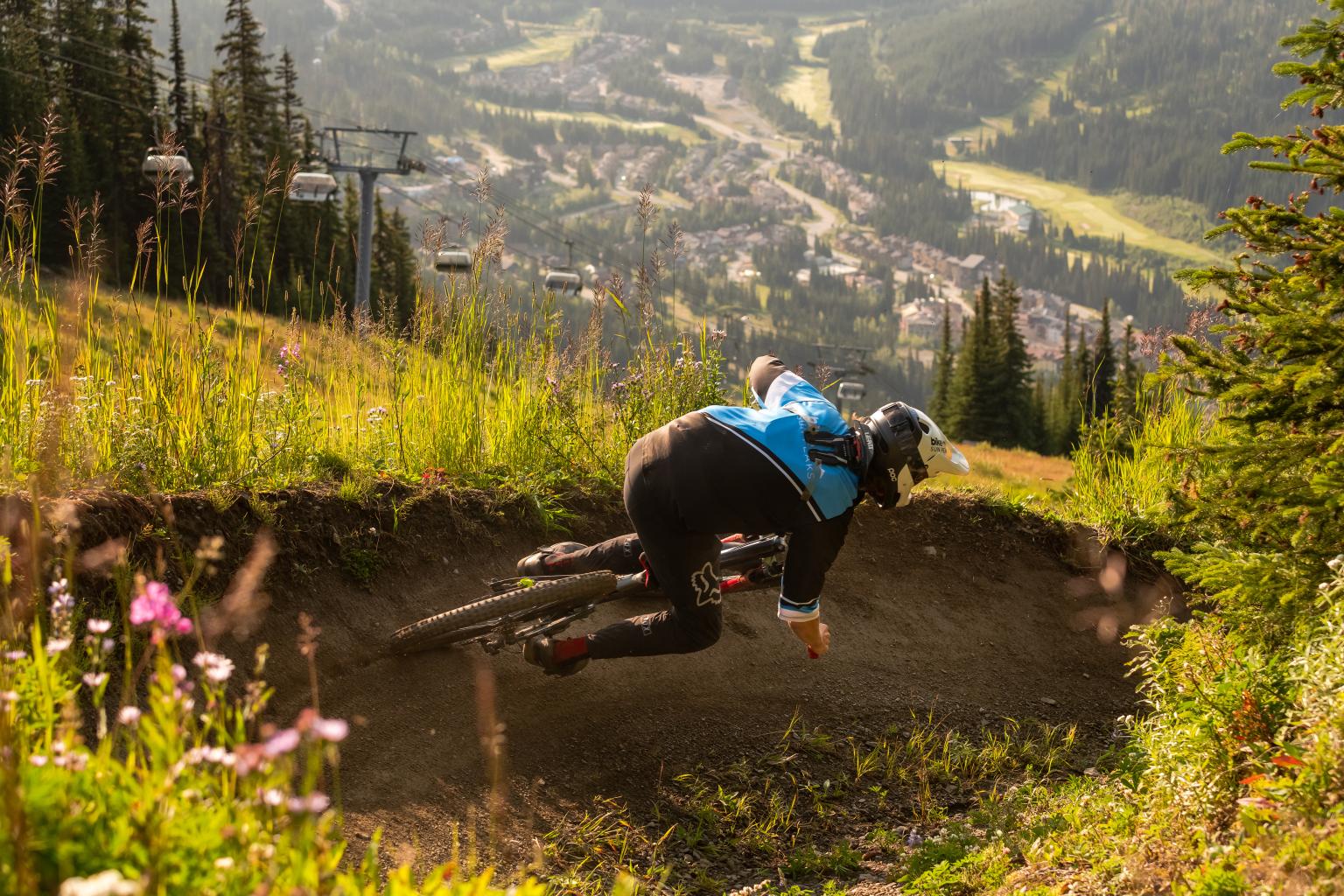 Mountain biker at Sun Peaks Resort riding a dirt trail amidst lush greenery, distant village in background.