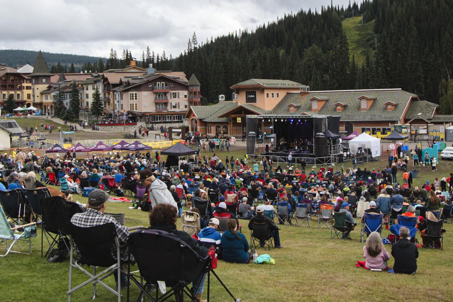 Outdoor concert with crowd sitting on grassy hillside, stage to the right, buildings in the background.