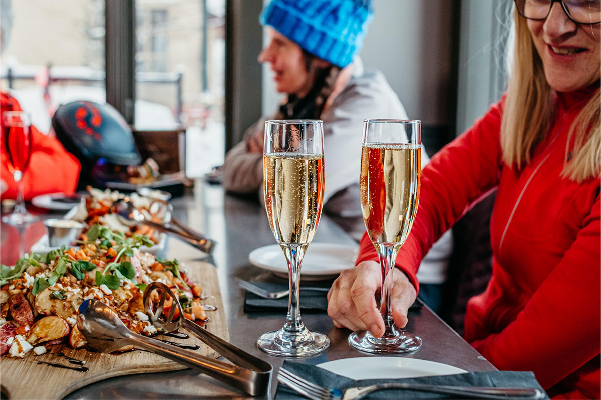 Two glasses of sparkling wine with a potato salad appetizer on a table, with two women in winter clothing in the background..