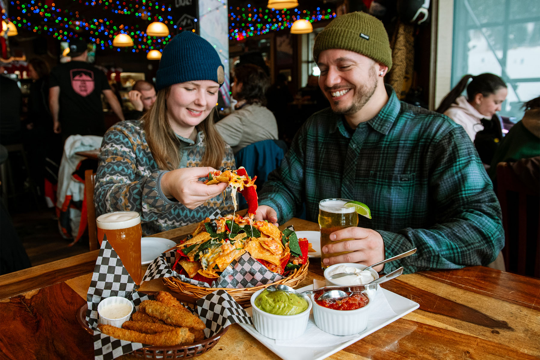 Woman pulls a chip from a plate of cheesy nachos and man smiles while enjoying apres at a bar.