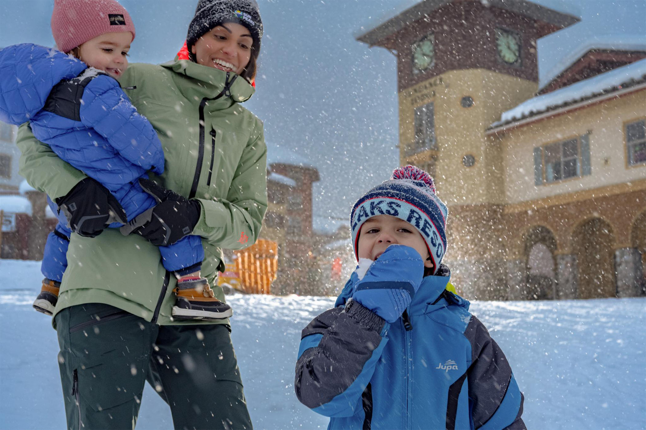 Little boy smiles and eats snow while his mother holds his little sister on her hip smiling.