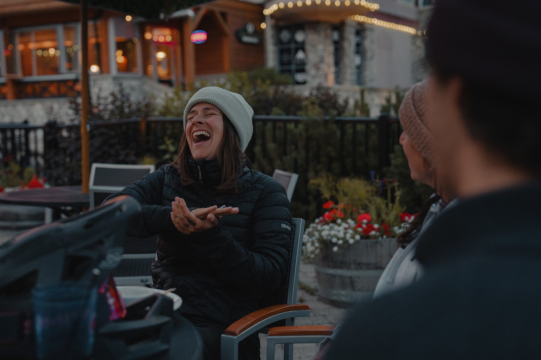 A woman laughs while sitting on a patio outside with two other people.