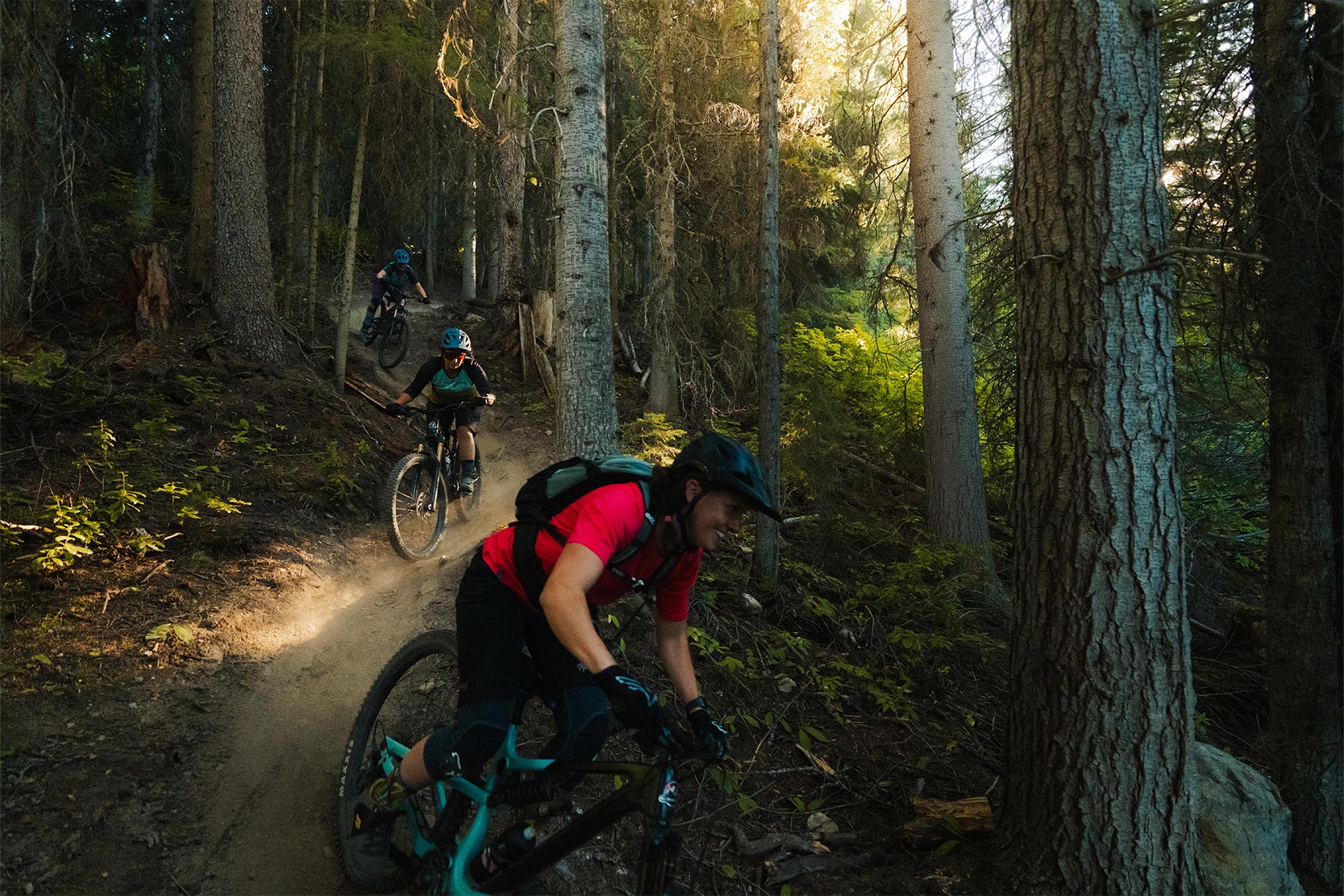 Three women riding their bikes down a trail with trees around them and sunlight seeping in