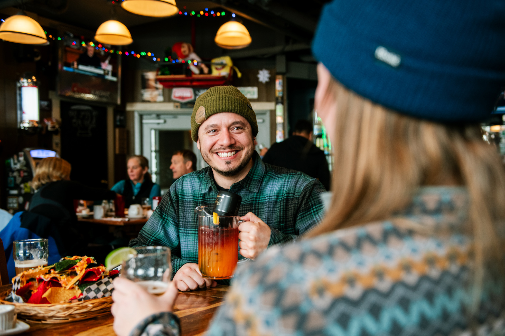 A man in a green hat holds a red and orange cocktail and smiles across the table full of nachos and drinks in Bottoms Bar & Grill.