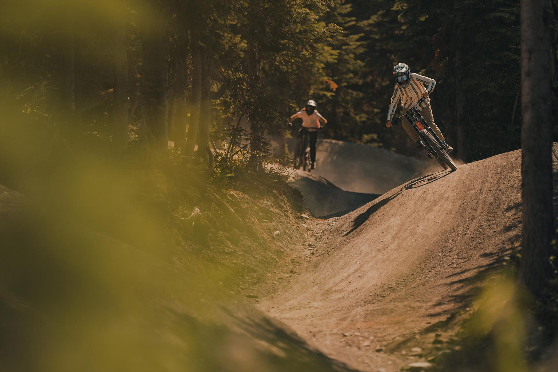 Two bikers coming down a rolling trail with greenery in foreground.