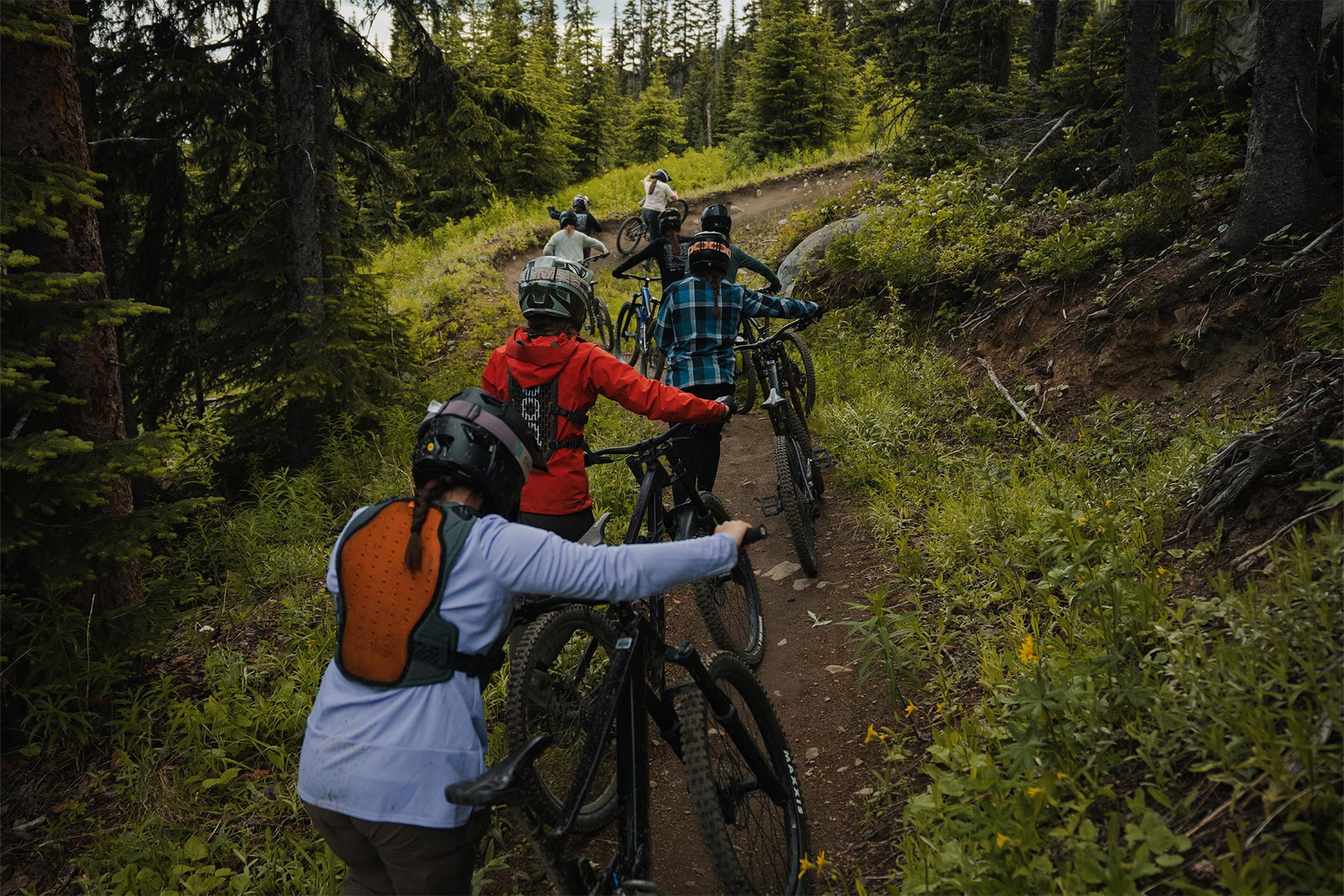 a group of ladies pushing their bikes up a dh trail with trees around them